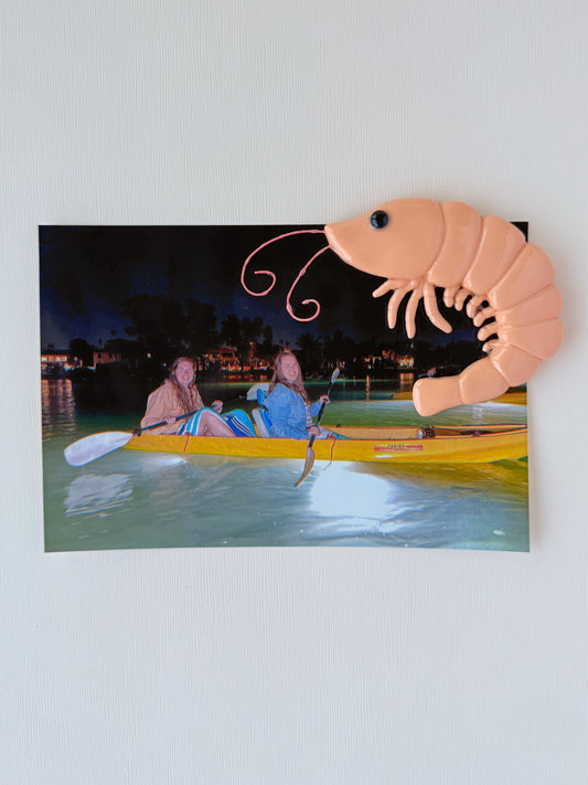 Photo of two people in a kayak on water with a large pink shrimp magnet with a white background